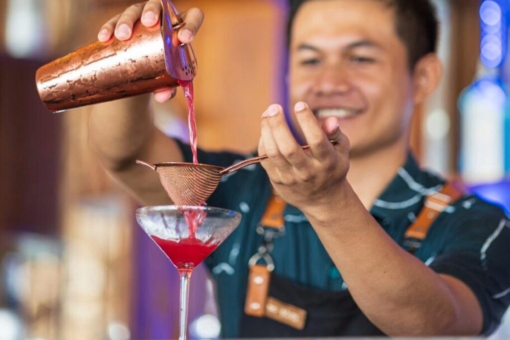 Bartender pouring a red cocktail through a strainer into a glass, preparing drinks at a beach club.