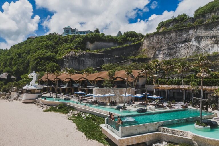 Beach club in Bali with large pools, oceanfront seating, and cliffs rising behind the resort area.