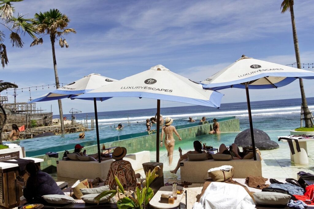 Beach club with infinity pool facing the ocean, guests relaxing under umbrellas and enjoying the seaside view.