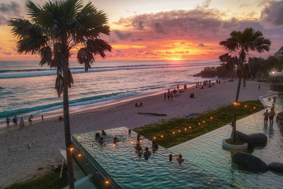 Beach club in Bali at sunset with ocean view, infinity pool, palm trees, and people relaxing by the beach.