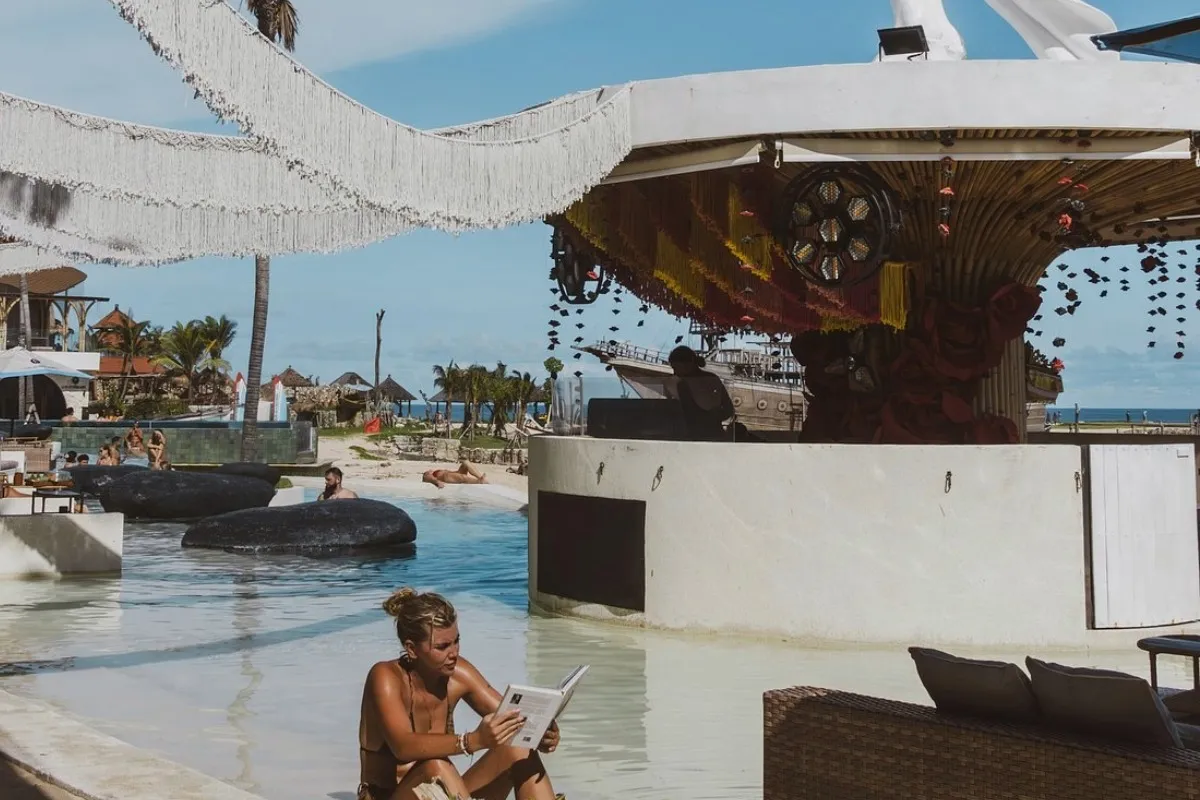 Woman reads by pool near bar, people relax and swim at a beach club in Bali.