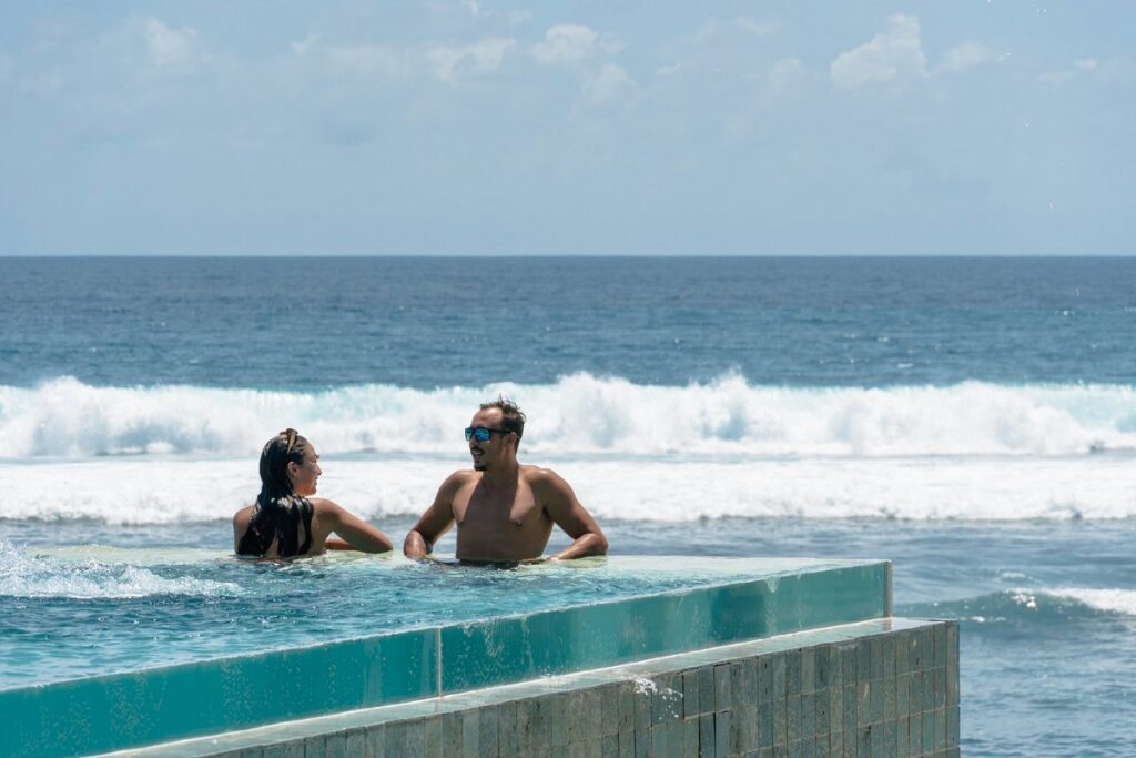 Couple relax in infinity pool facing ocean waves, showing beach club experience in Bali.