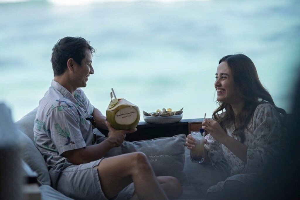 Couple relaxing by the ocean, drinking cocktails and coconut, enjoying a memorable beach club dining experience.