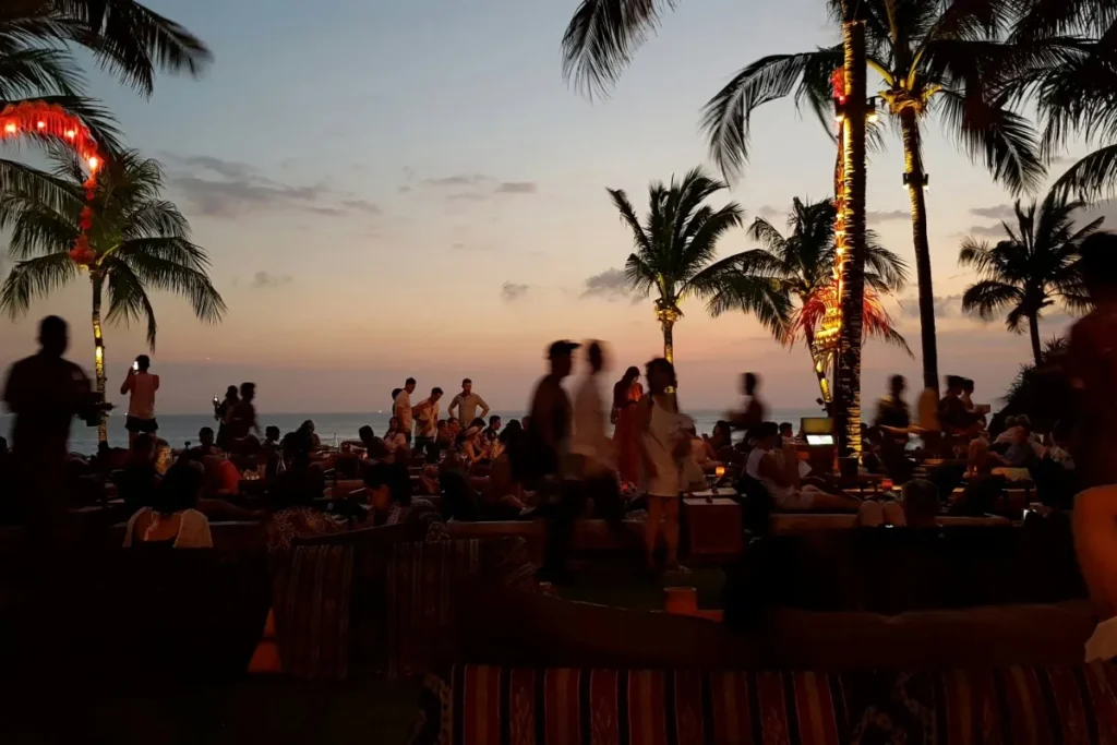 Crowded beach club at sunset with palm trees and lights, showing nightlife and social scene in Seminyak.