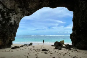 A traveler stands on a quiet South Bali beach framed by cliffs, facing clear ocean waves and open sky.
