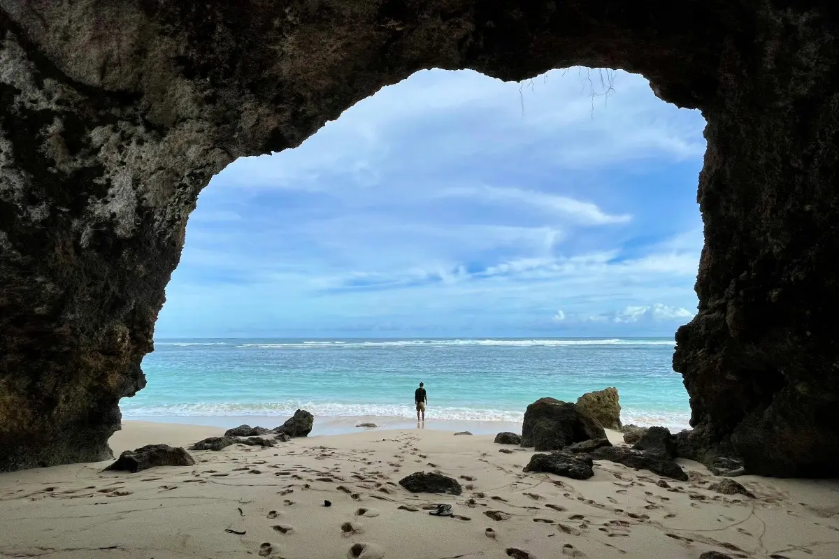 A traveler stands on a quiet South Bali beach framed by cliffs, facing clear ocean waves and open sky.